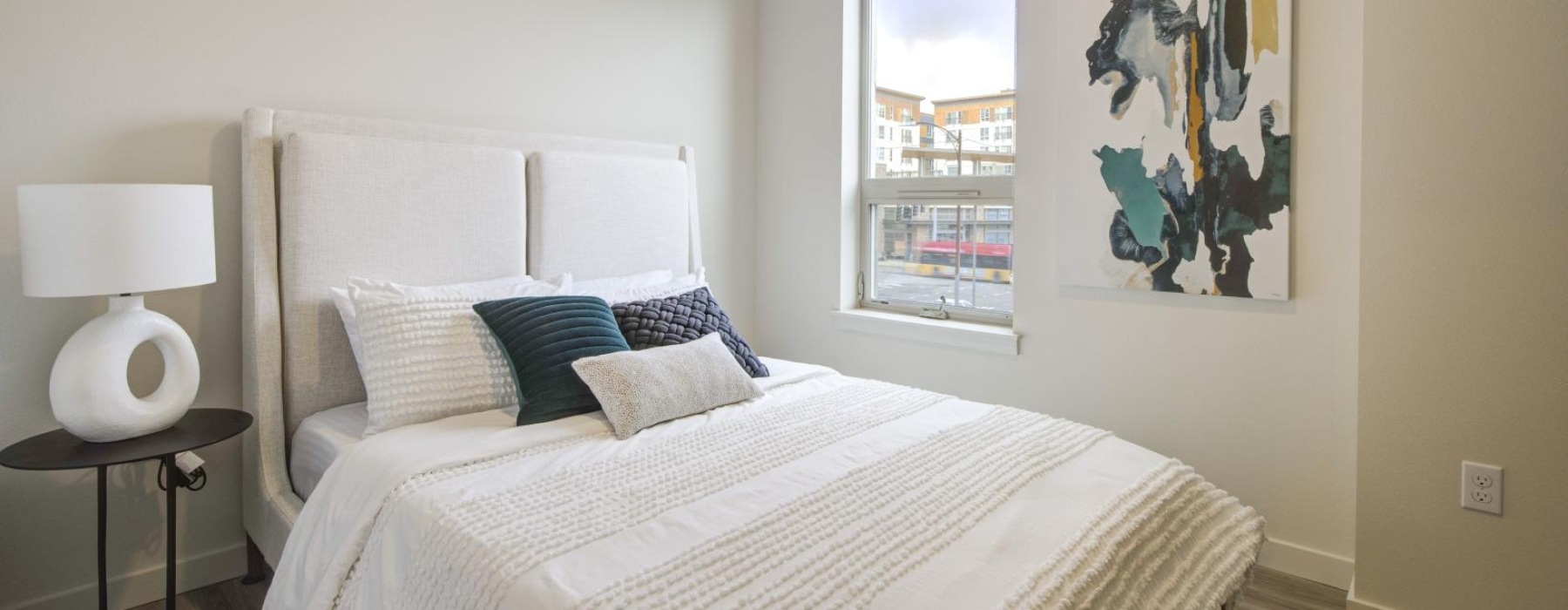 Bright apartment bedroom with a white bed, teal accent pillows, abstract wall art, and a street-view window.