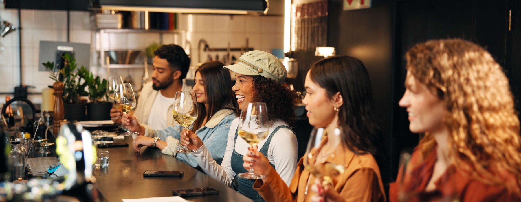 Five friends laughing and holding glasses of white wine at a bar counter.