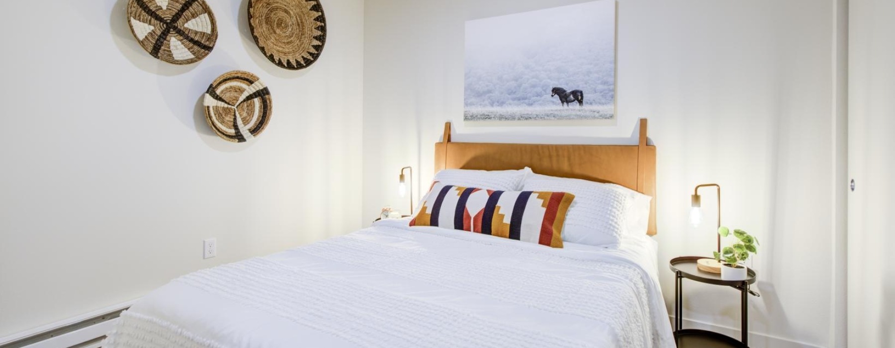 Bright apartment bedroom with a white bed, leather headboard, woven wall baskets, and a framed horse photo.